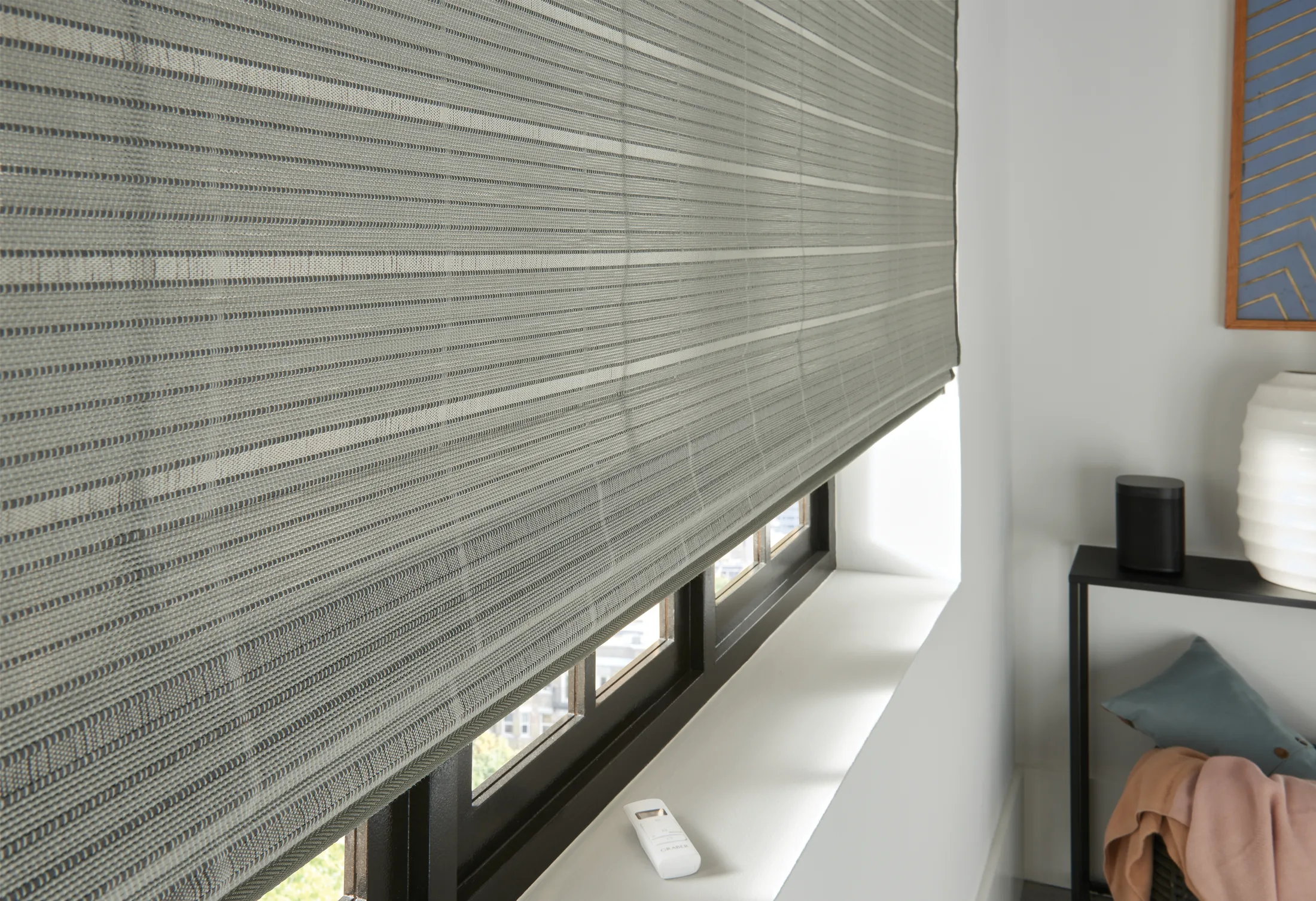A close-up shot of light gray woven wood shades. The shades are lowered most of the way to cover a large window. The room’s walls are white, and part of a black table can be seen to the right of the window.