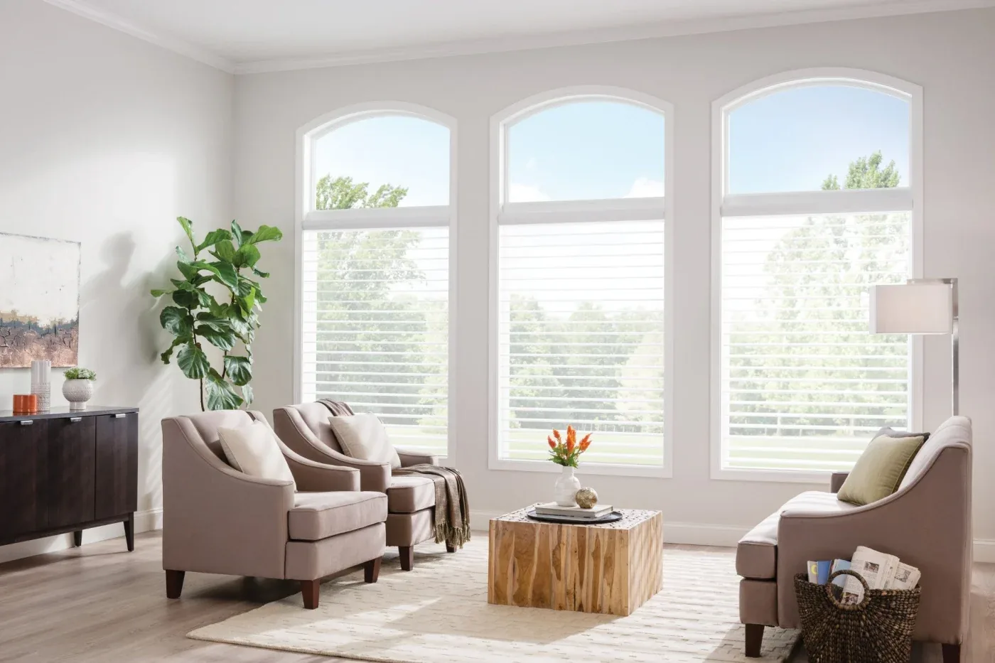 A bright living room with three tall arched windows, white blinds, soft neutral seating, a wood coffee table, and light modern décor