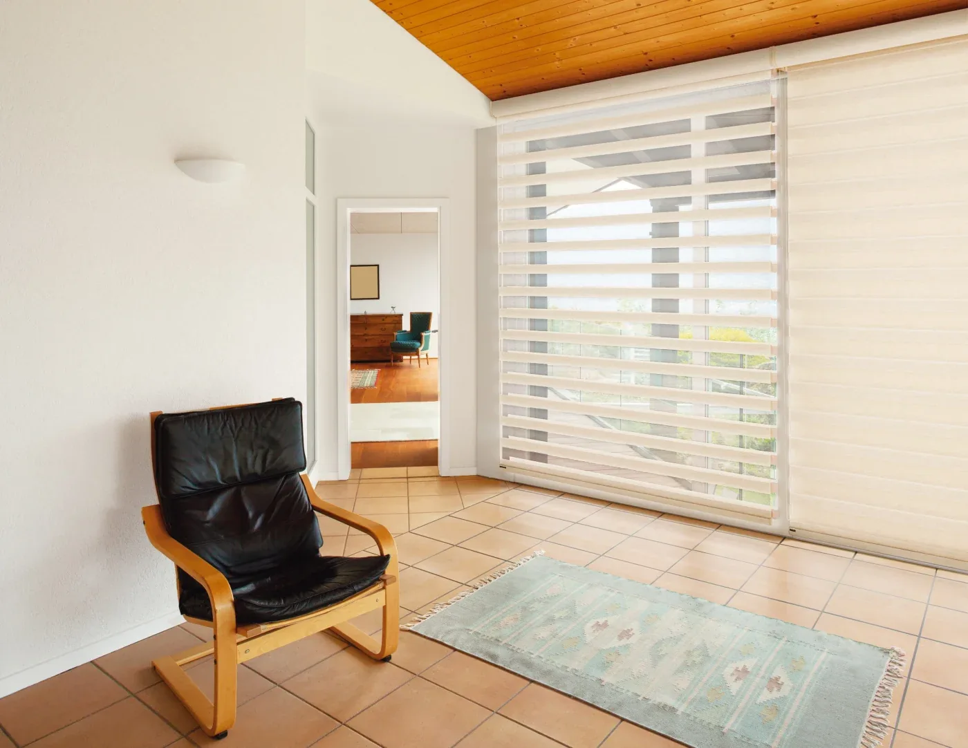 A bright room with layered zebra shades over tall windows, a black leather chair, tan tile floor, wood ceiling, and a light blue accent rug.
