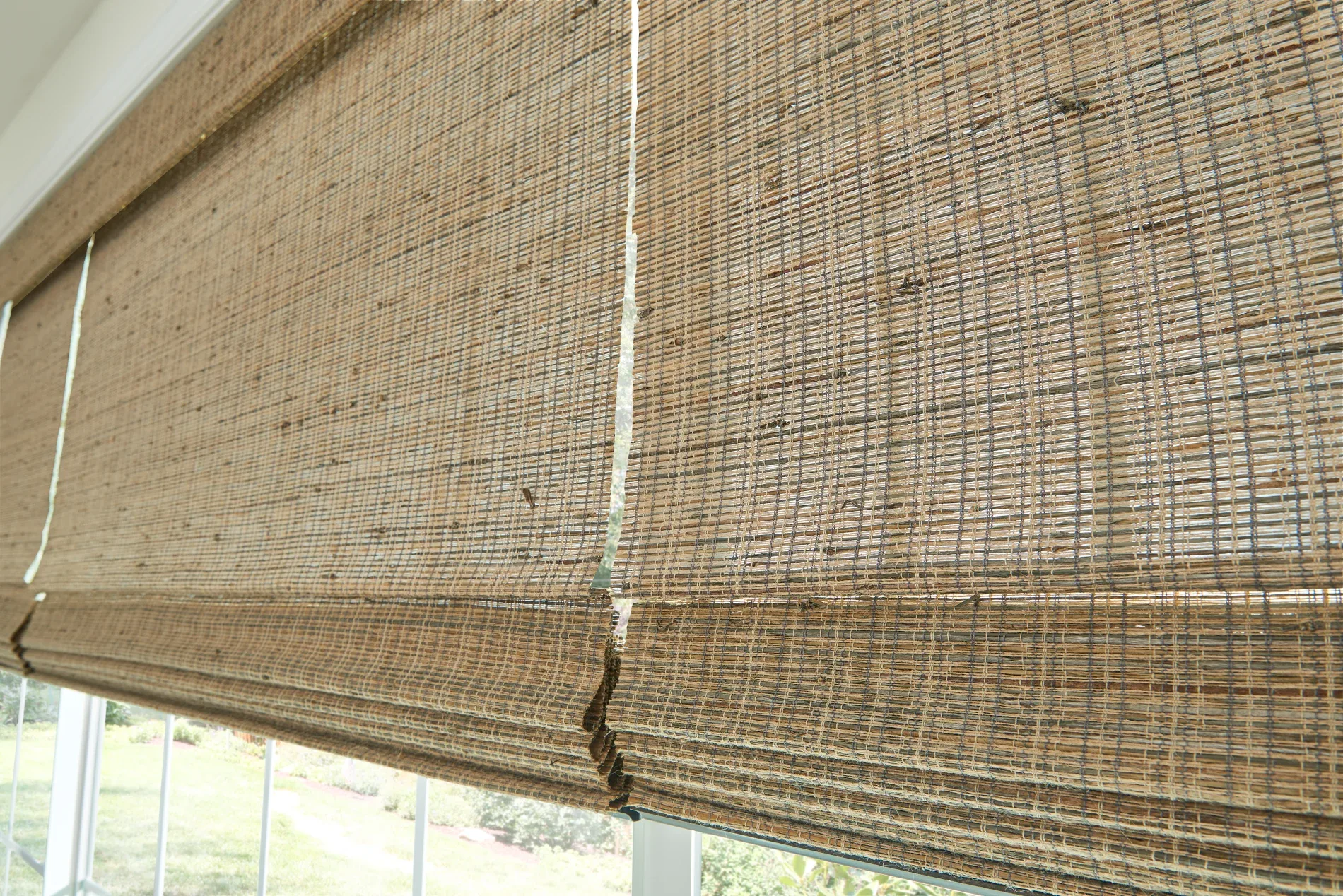 A close-up of three partially closed woven wood shades in a medium brown color. The shades are letting a moderate amount of light through, and beneath them, you can see out and into the yard on a sunny day.