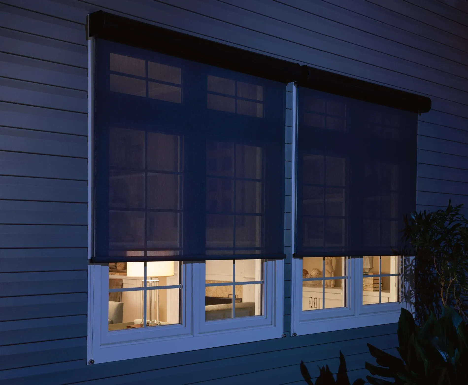 A close-up view of a home’s exterior at night. Two large windows are visible, with dark-colored motorized shades lowered almost all the way. There are lights on inside the house, and part of a thoroughly furnished living room is visible through the windows