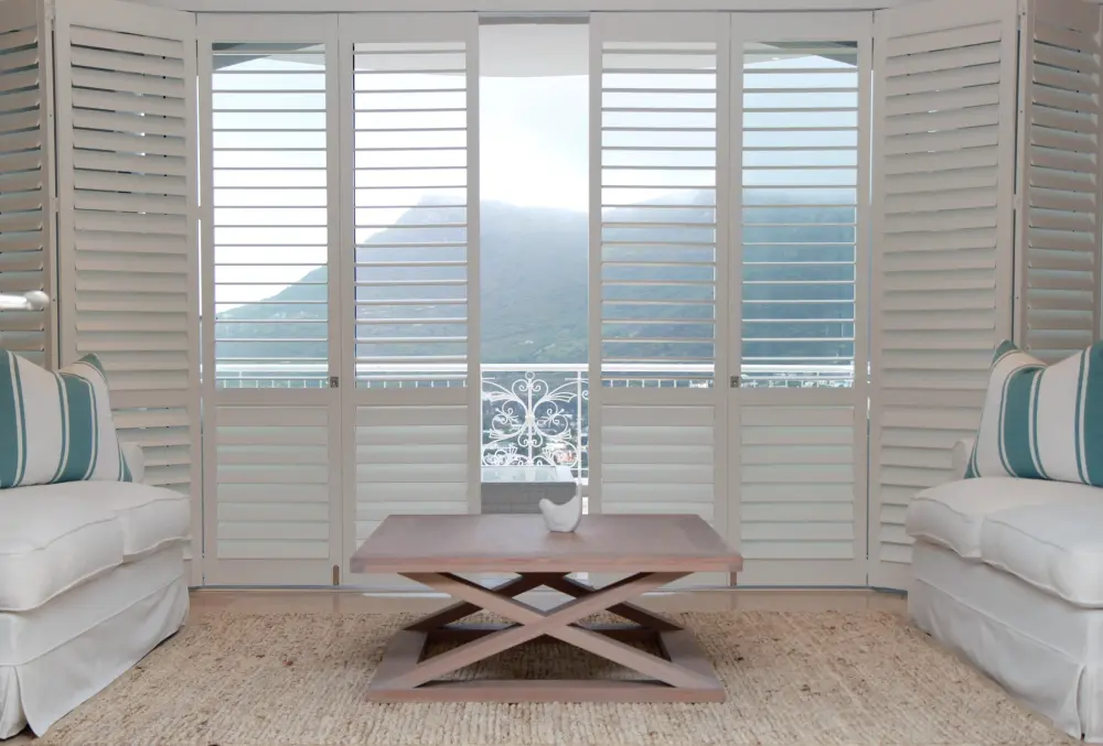 White plantation shutters frame balcony doors with mountain views, white sofas, striped pillows, woven rug, and wood coffee table.