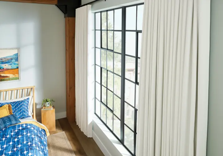 A bedroom with cream floor-length drapery, black-framed picture window, wood accents, and blue patterned bedding.