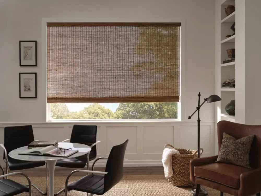 A home office with a woven Roman shade over a wide window, black chairs around a round table, a leather armchair, and built-in shelving.