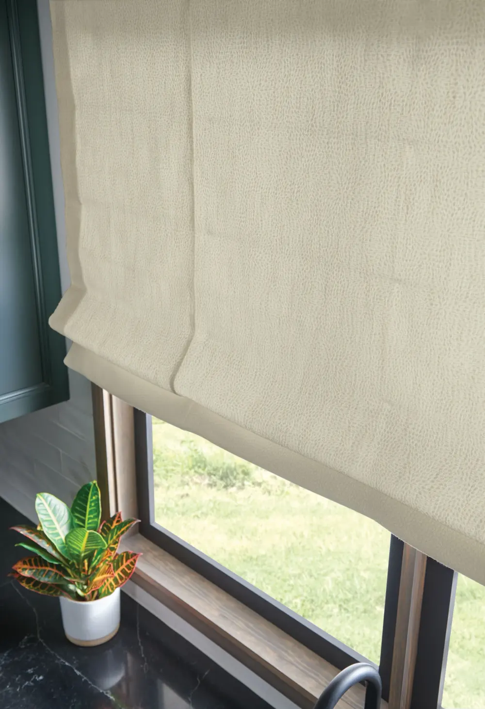 A close-up of a custom Roman shade in textured neutral fabric partially lowered over a window.