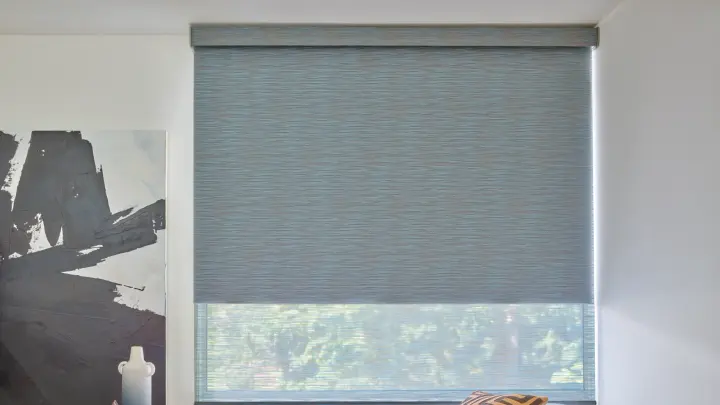 A blue-gray roller window shade is partly lowered in a modern room beside abstract wall art and minimalist décor.