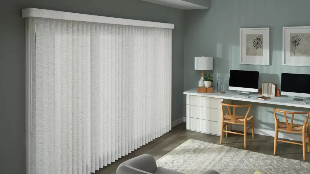 A home office with light gray vertical blinds, a white desk, two computers, wood chairs, framed wall art, and a soft area rug.