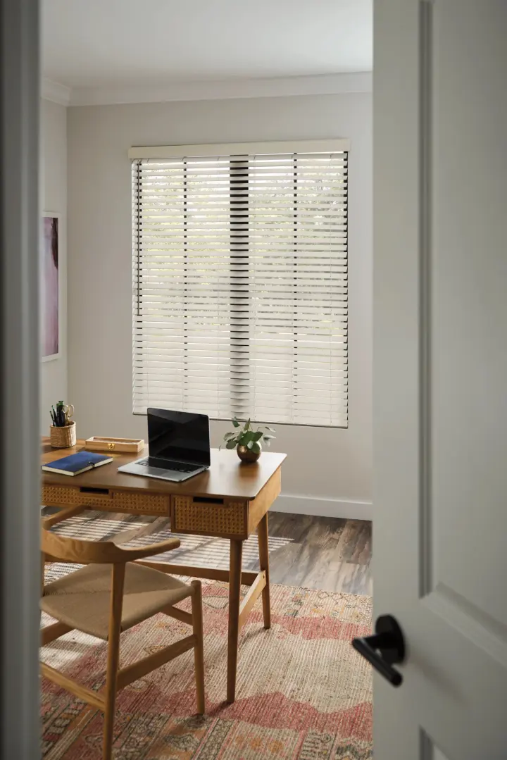 A view into a home office with a brown wooden desk and chair in the center of the room. The walls and ceiling are white, and the flooring is dark brown and gray wood grain, with a bright area rug on top of it. There is a large window on the back wall, covered in white horizontal blinds.