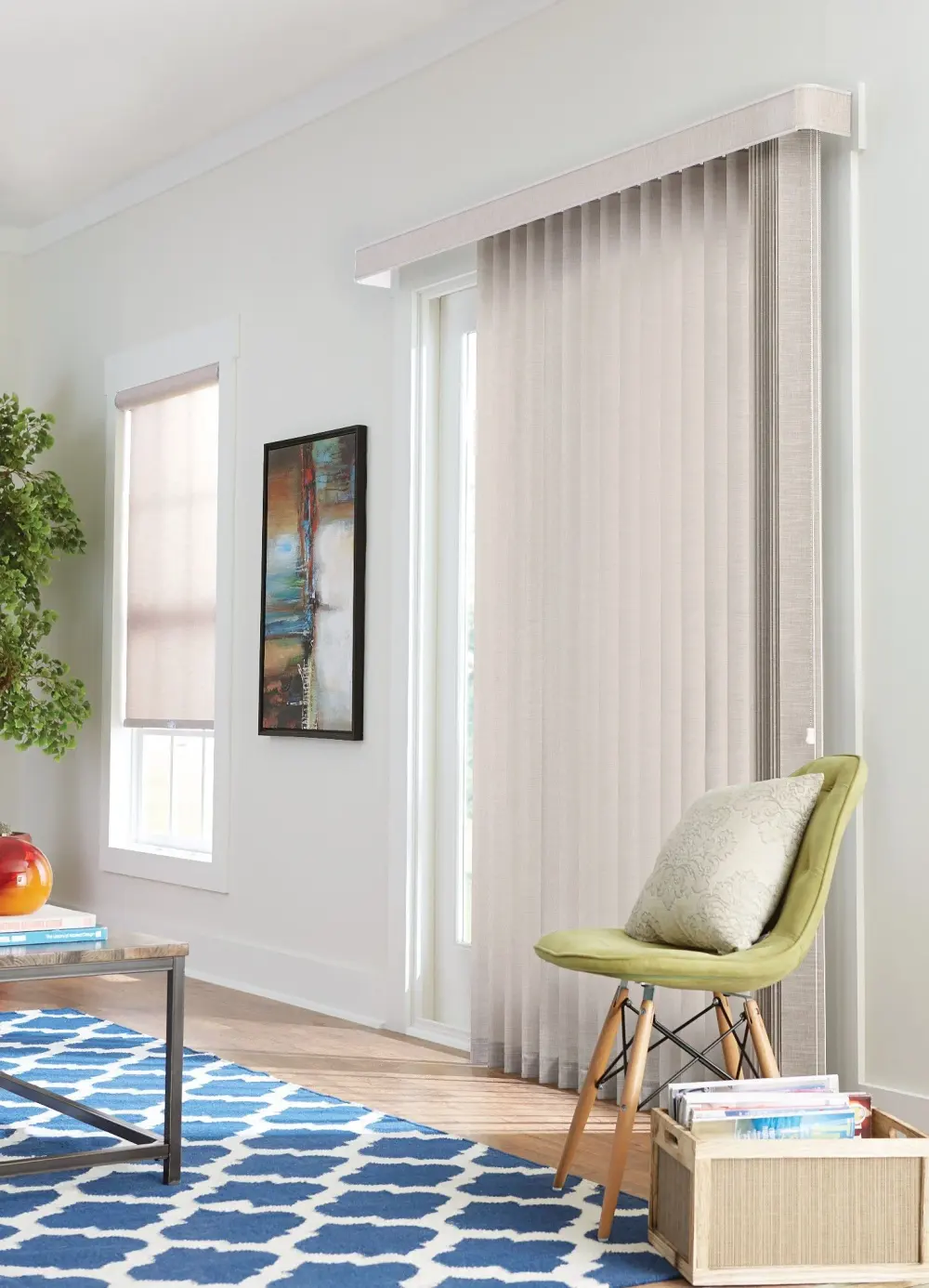 A living room with light beige vertical blinds, a green accent chair, a blue patterned rug, wall art, and a tall window shade.