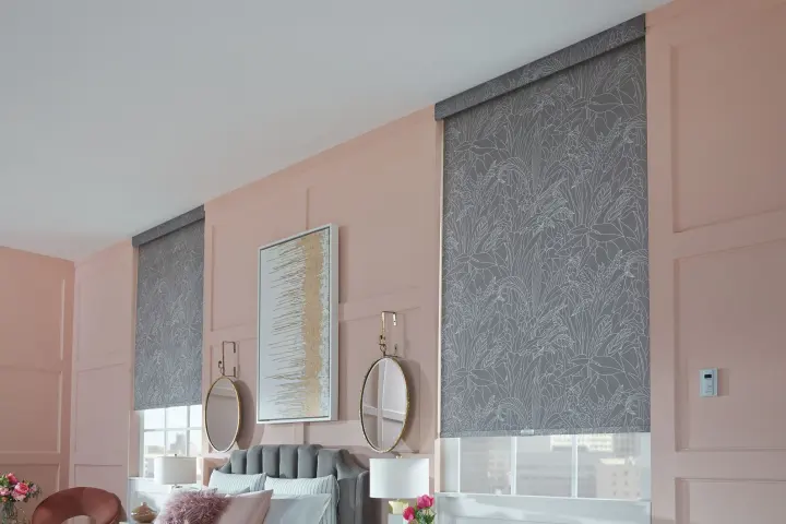 A pair of patterned roller shades covers the windows in a room with pale pink walls. The shades are dark gray and have a white floral pattern on them. There is a plush gray headboard against the wall between the windows.