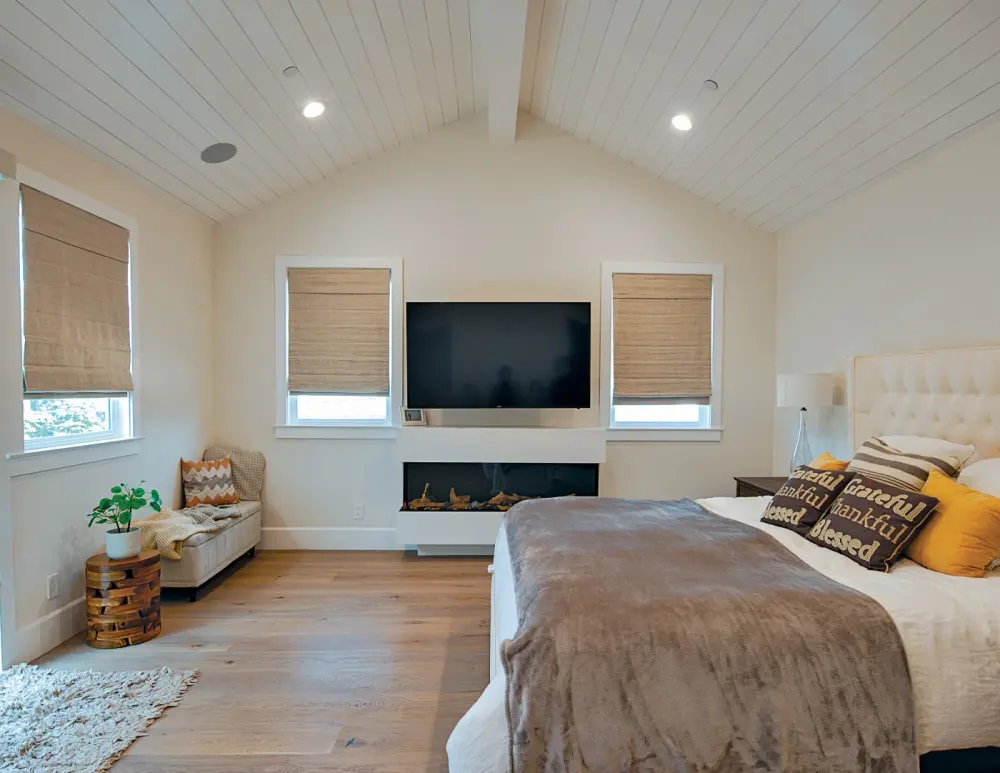 A bedroom with a TV mounted on the far wall and brown motorized shades covering the windows.