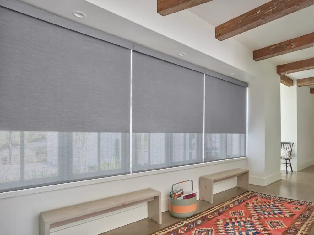 A large gray roller window shades in a modern room with exposed wood ceiling beams, simple bench seating, and a colorful patterned rug.