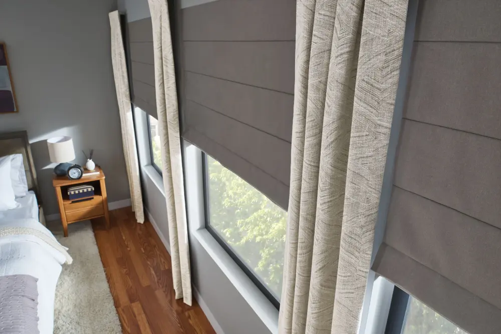 A view of a set of windows with dark gray shades, and long, patterned gray and white drapes. Part of a bed is visible on the left, and there is a small wooden nightstand between the bed and the windows.