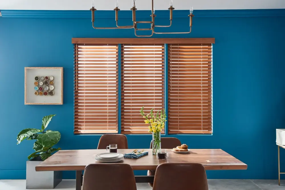 A dining room with rich wood blinds, a bold blue accent wall, wood table, brown chairs, brass chandelier, and floral centerpiece.