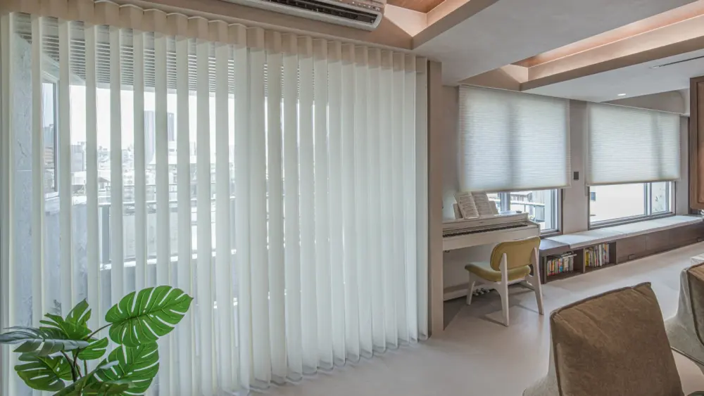 Home with vertical drapes and cellular shades on windows with piano setup and small wooden bench with books underneath it