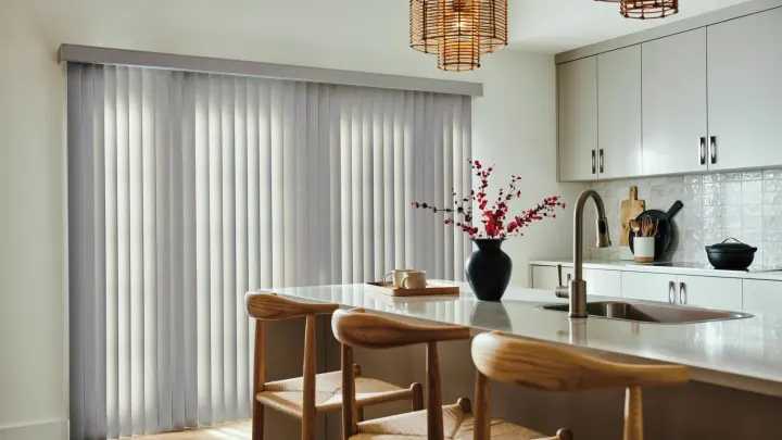 A modern kitchen with light gray vertical blinds, white cabinets, island seating, pendant lights, and a black vase centerpiece.