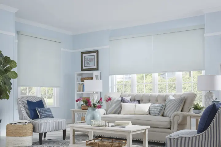 A living room with light blue walls and white roller shades over the windows.