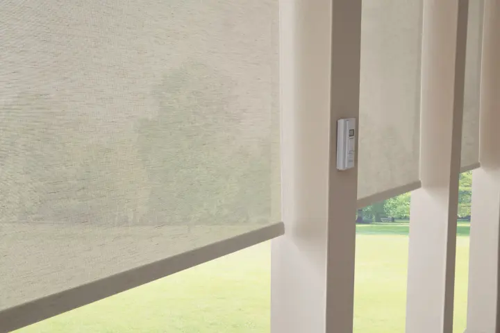 A close-up view of a pair of large windows with semi-opaque roller shades covering them. The shades are cream colored, and they are halfway closed, covering the top of the windows and showing off green grass toward the bottom.
