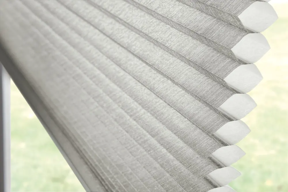 Up close view of gray cellular window shades. They are pulled partially down, covering the top of the window, though they are open enough to show part of the yard through the bottom of the window.