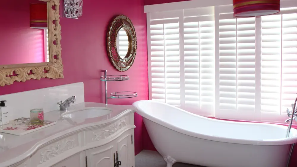 Bathroom with classic design and red walls with tall white shutters on both windows behind the bathtub