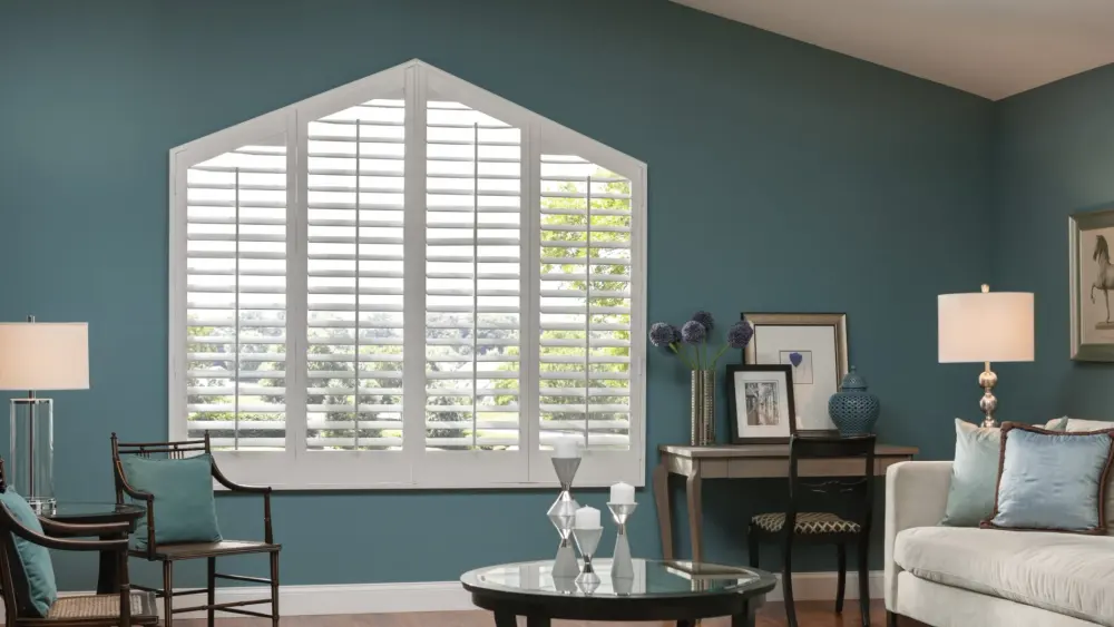 A living room with white gabled interior shutters, teal walls, cream sofa, wood chairs, desk, and glass coffee table.