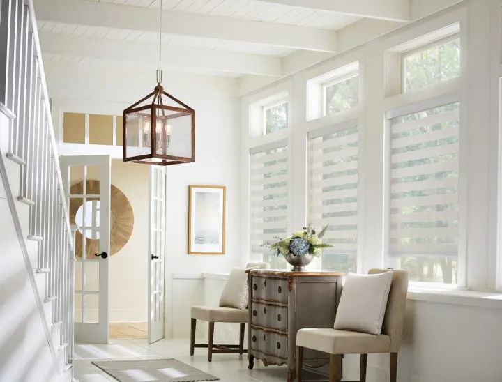 A bright entryway with layered zebra shades, tall windows, white staircase, lantern pendant light, and a small accent table with two chairs.