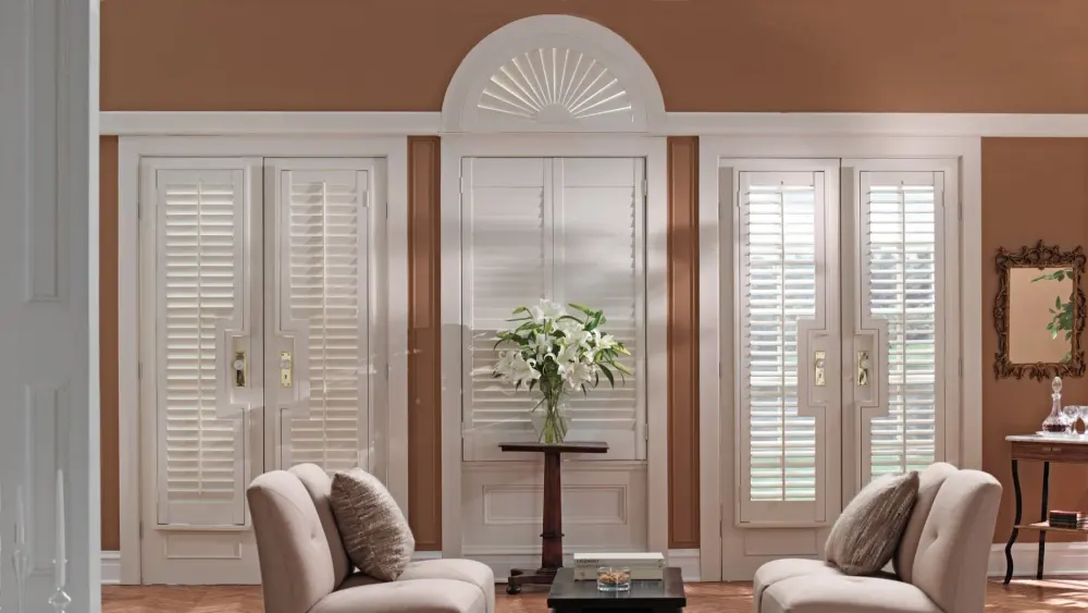 Elegant sitting room with white interior shutters, arched transom shutter, upholstered chairs, side tables, and a vase of white lilies.