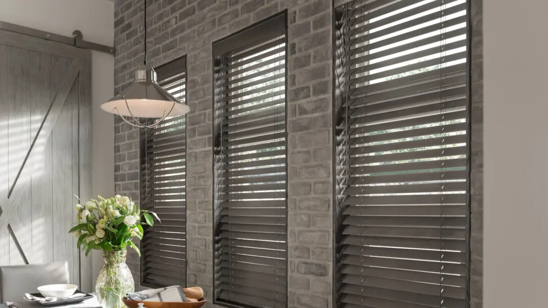 Dining area with dark wood blinds on tall windows, gray brick accent wall, pendant light, and floral table centerpiece.