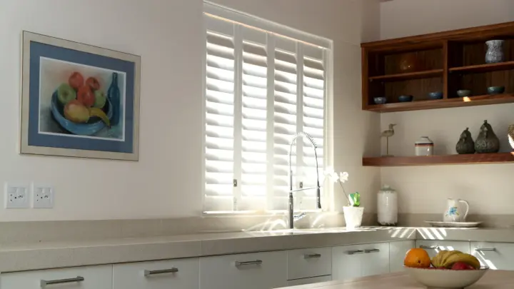 Kitchen space with a white interior, kitchen counter with beige countertop, brown shelving near white wall, and white half-closed shutters
