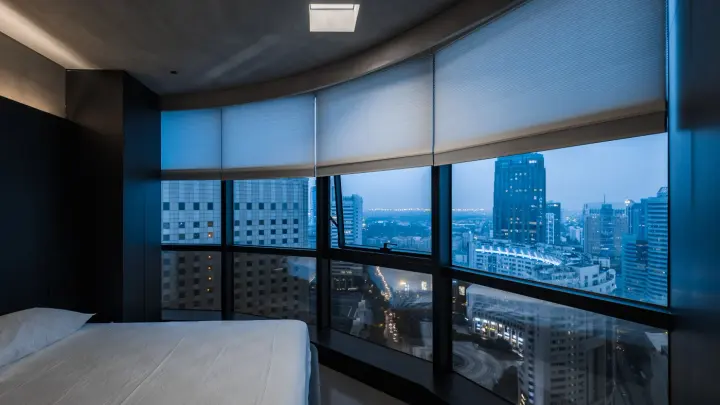 A bedroom with a wall of windows with motorized shades open overlooking the city