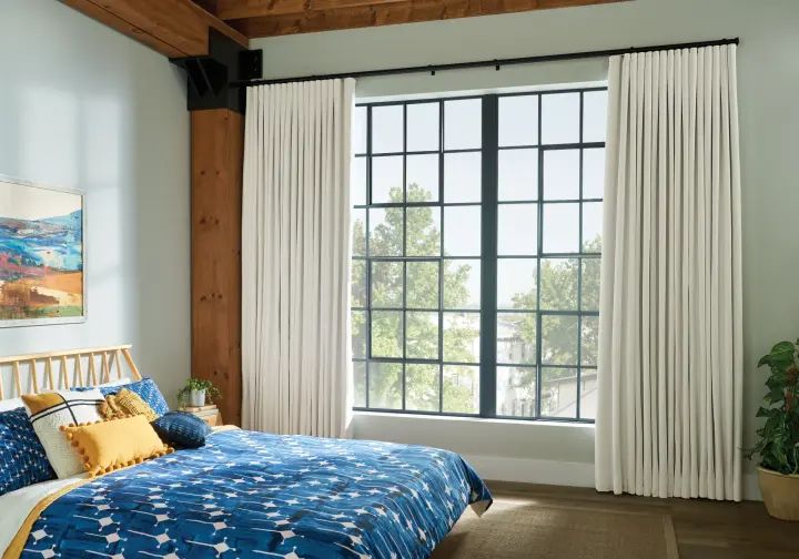A wide shot of a bedroom featuring a large, wide bed with blue and yellow patterned bedding. A large window with multiple square panes and a black frame takes up the majority of the far wall. The window shades are long and white, and are opened to leave a space in the middle for light.