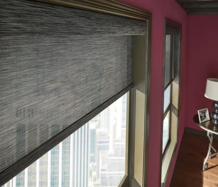 A view of the top portion of two windows with gray roller shades. The shades are halfway closed to partially block the light entering the room. The room's walls are dark pink, and there are shadows on the wooden floors.