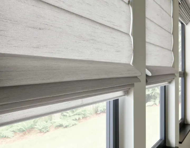 A close-up of light gray Roman shades on a pair of windows with black frames. The shades are opaque, but light is still visible through them.