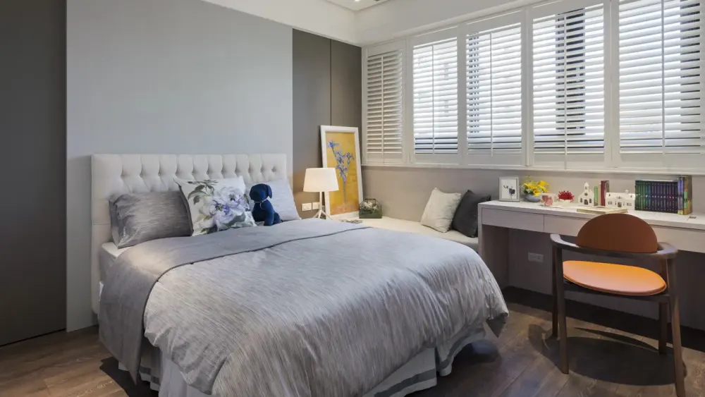 Bedroom with light grey interior, bed with grey blanket, reading desk with books, brown chair, and white half-closed wood shutters