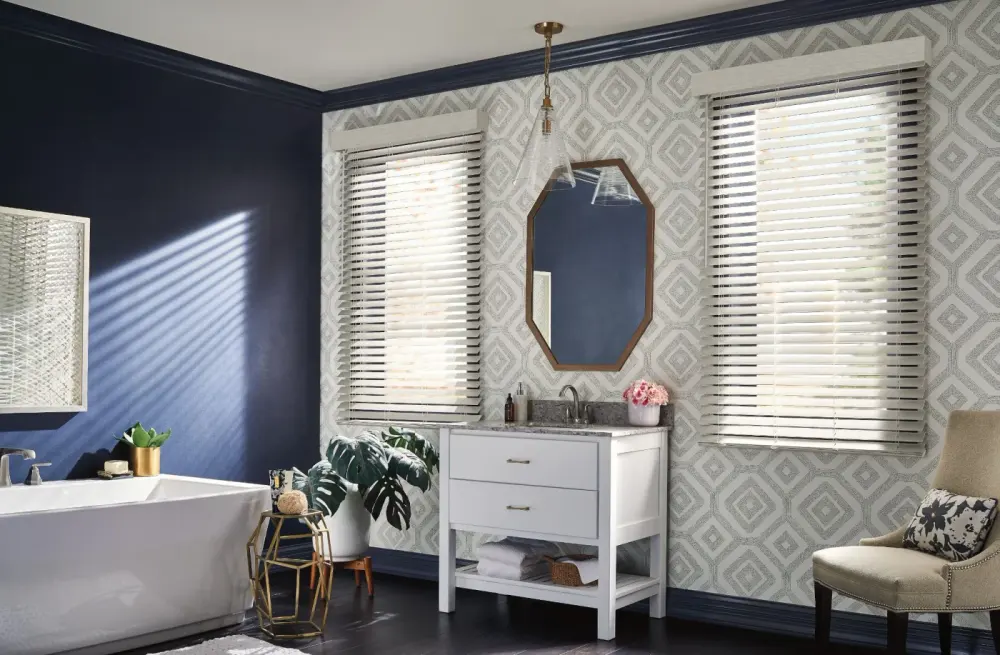 A bathroom with a standalone tub in front of a blue wall and two windows with blinds.