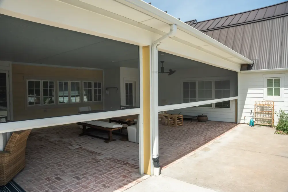 A pair of patio roller shades is partially pulled down to cover large outdoor openings. The shades are mounted on the outer edge of a patio's roof. The home has white and yellow siding, and the covered portion of the patio is red brick with white and wicker furniture.