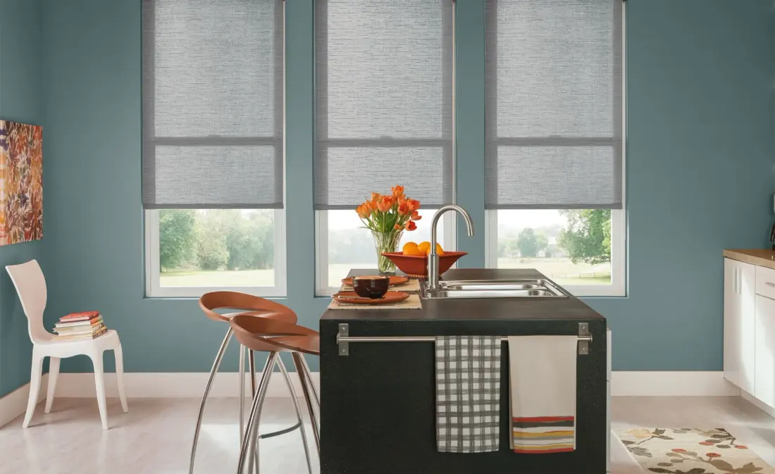 Modern kitchen with three gray roller window shades, teal walls, black island sink, bar stools, orange flowers, fruit bowl, and bright outdoor views.