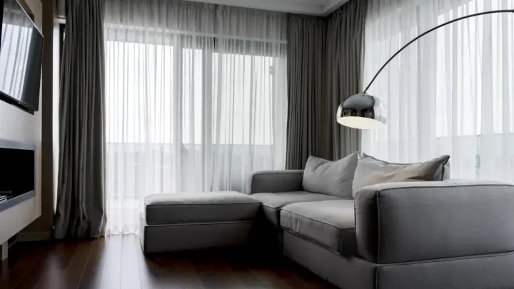 A living room with two walls of windows covered by layered drapery. The bottom layer is sheer white panels, and the top layer is dark gray.