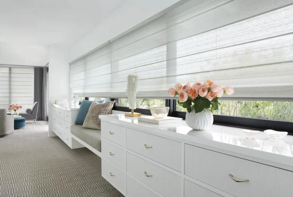 A bedroom with two matching white dressers and a small bench seat between them underneath a large window covered by custom window shades.
