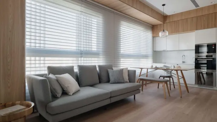 Beautiful living room with light-colored hardwood floors, gray couch, white cabinets, black appliances, and gray window shades