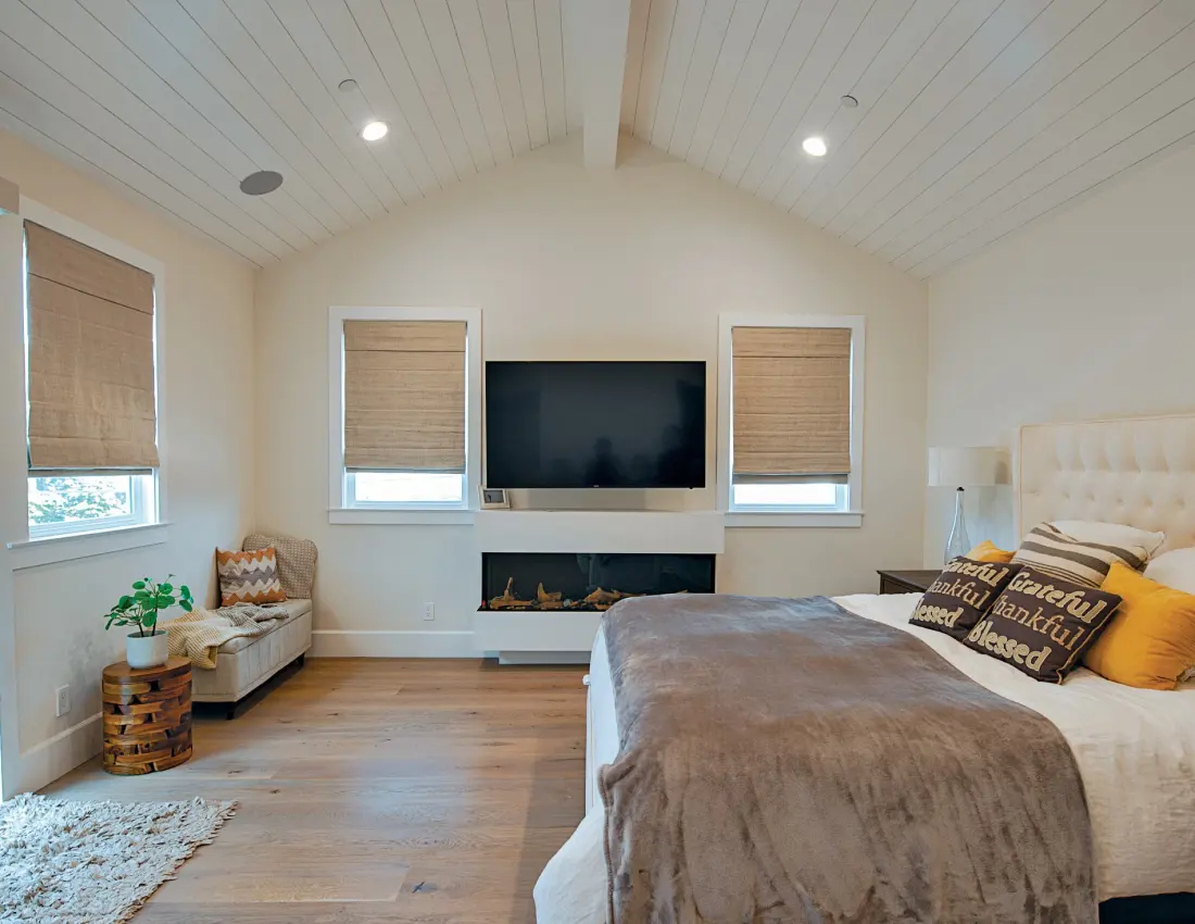 Bright modern bedroom with sophisticated Roman shades, upholstered bed, fireplace, TV, accent chair, wood floors, and vaulted shiplap ceiling.
