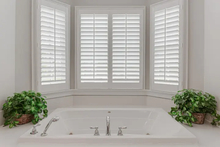 White plantation shutters and soft plant accents over a clean, modern soaking tub