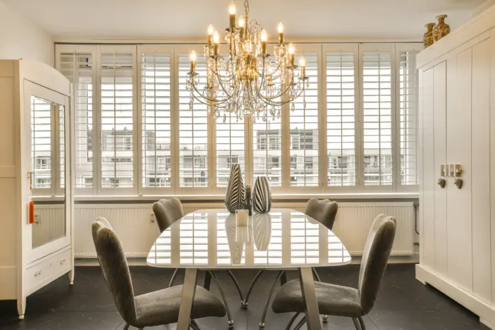 An elegant dining room with a table, chairs, chandelier, and white shutters over the windows.