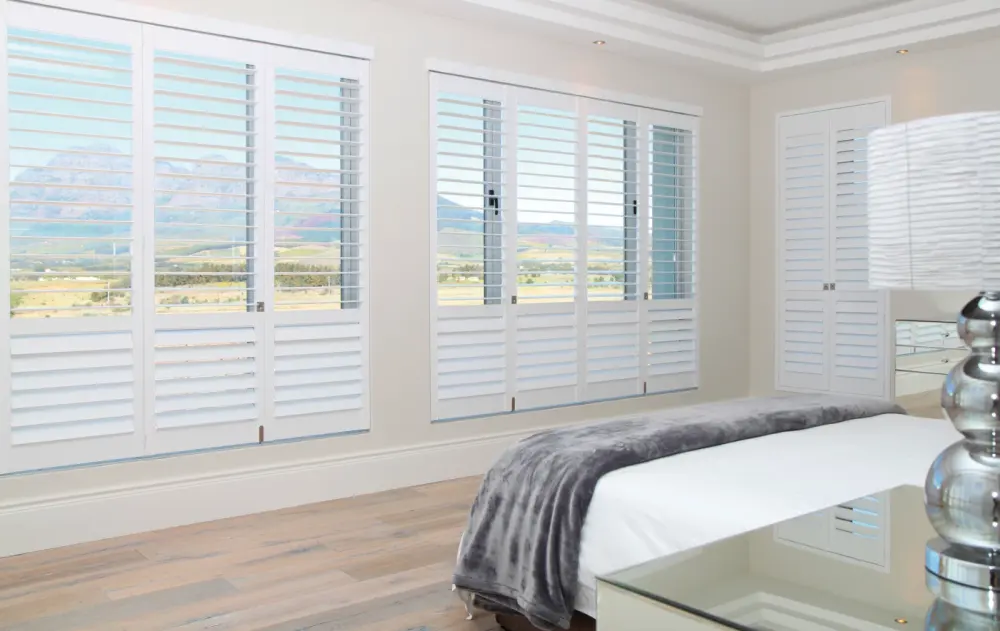 A bedroom with a wall of windows covered with white indoor shutters.