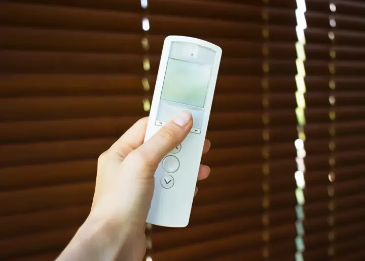 Hand holding a remote control with window shades in background