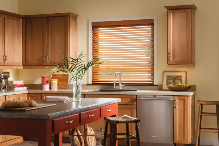 A kitchen with wood cabinets, stainless steel dishwasher, butcher-style island, bar stools, and wood blinds over the sink window.