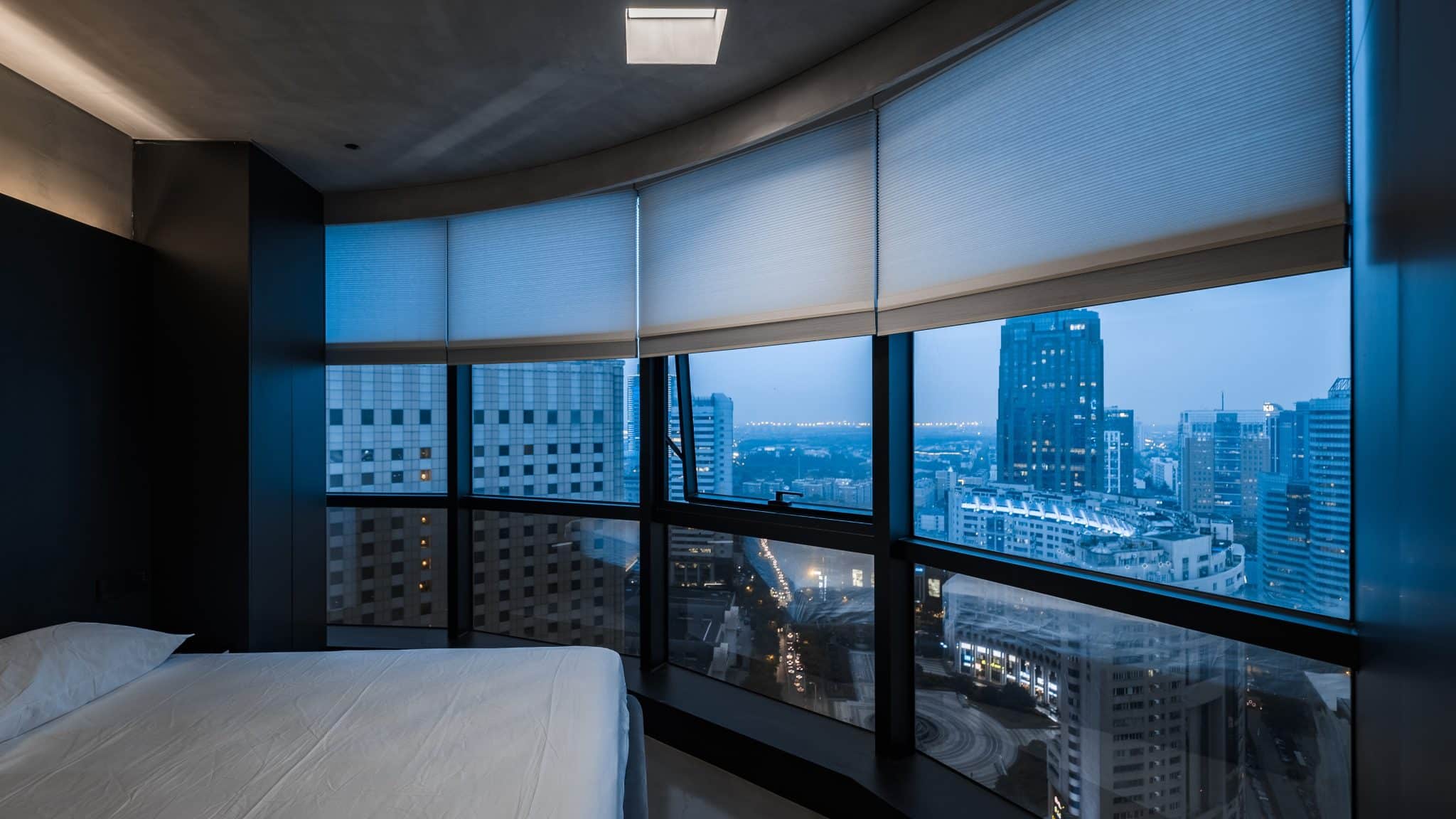 A Bedroom With A Wall Of Windows With Motorized Shades Open Overlooking The City