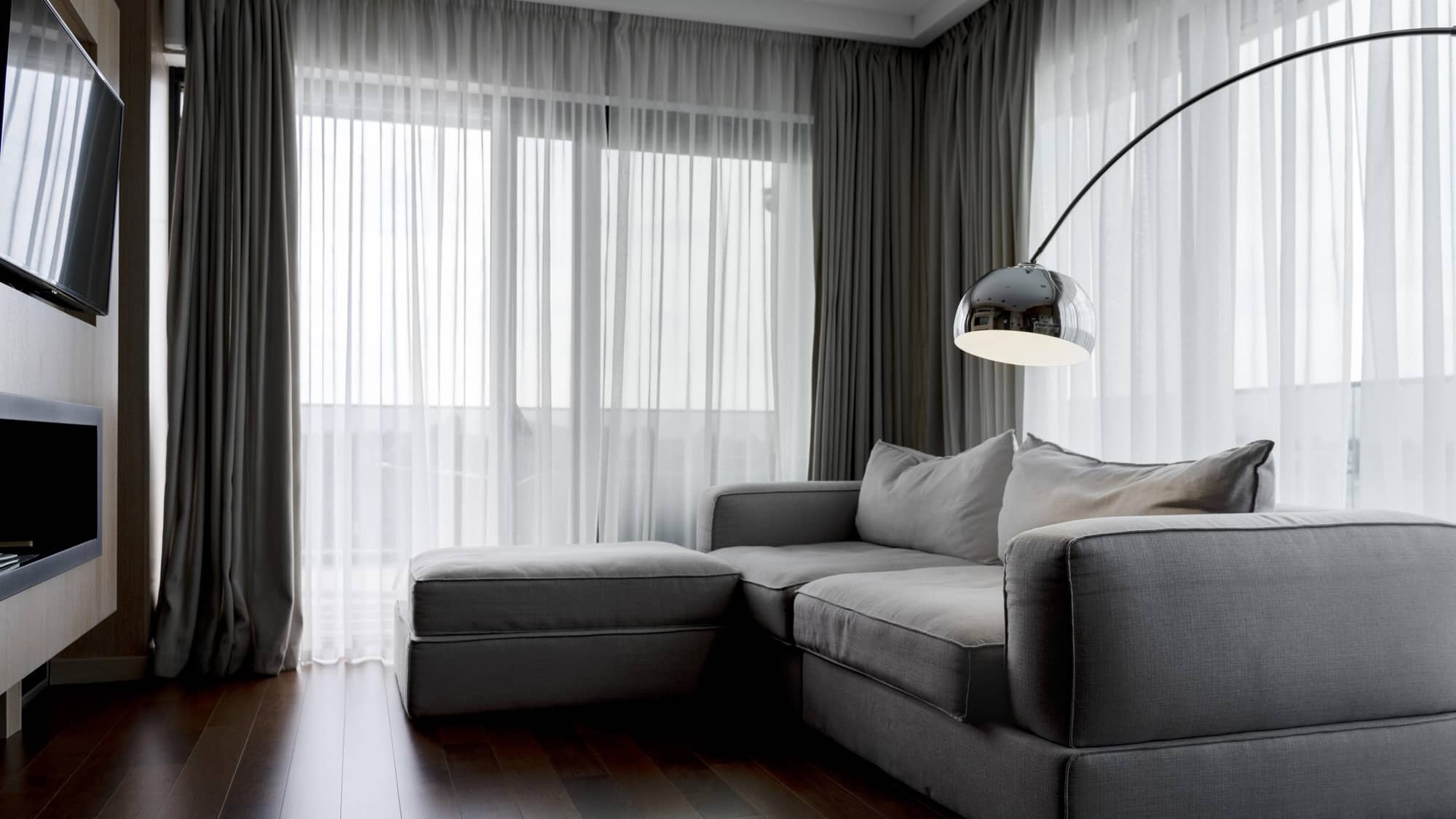 A Living Room With Two Walls Of Windows Covered By Layered Curtains. The Bottom Layer Is Sheer White Panels, And The Top Layer Is Dark Gray.