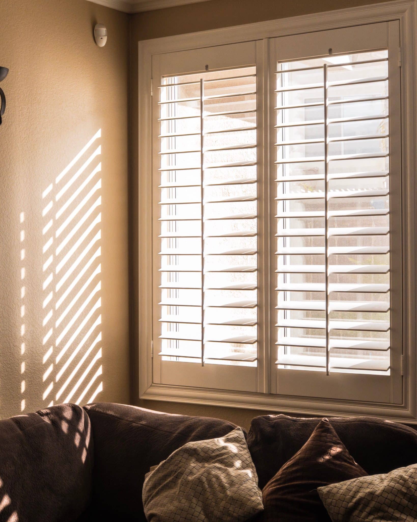 White Shutters Installed On A Window In The Living Room Area, With The Edge Of The Couch Near The Window.